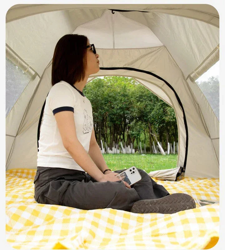 Woman sitting inside beige camping tent on yellow checkered blanket holding smartphone, view of green trees outside