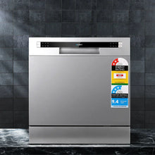 Stainless steel benchtop dishwasher with energy and water rating stickers on dark tiled background