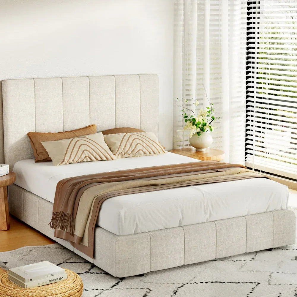 Modern queen upholstered bed with beige fabric, brown accent pillows, and a cozy blanket in a bright bedroom.