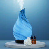 Blue glass aroma diffuser emitting mist, with essential oil bottles and white flowers