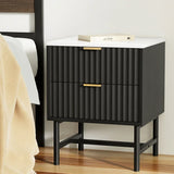 Black bedside table with ribbed drawers, gold handles, and white top beside a bed