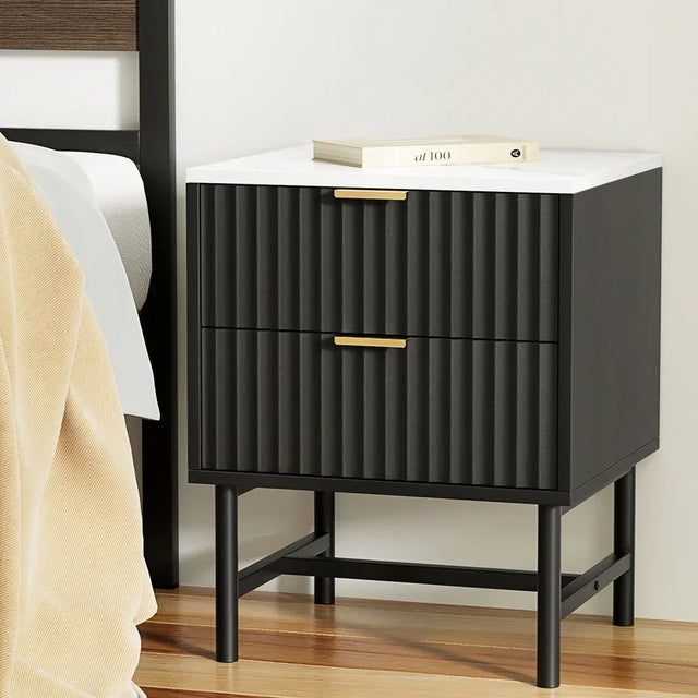 Black bedside table with ribbed drawers, gold handles, and white top beside a bed