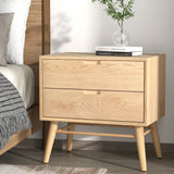 Light wood bedside table with two drawers, angled legs, books and a vase on top, bedroom setting