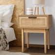 Modern bedside table with natural wood finish, cane drawer front, lamp, and striped cup