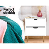 Modern white bedside table with two drawers beside a bed, featuring a book and pink vase.