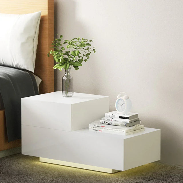 Modern white bedside table with LED lighting, alarm clock, vase, and books in a bedroom setting