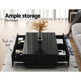 Modern black coffee table with glass top, four push-to-open storage drawers, and vase decor.