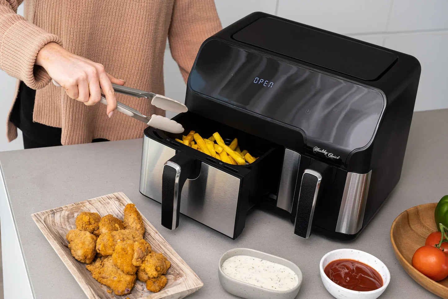 Person using stainless steel tongs to take fries from a black and silver air fryer with fried chicken and dipping sauces on the counter