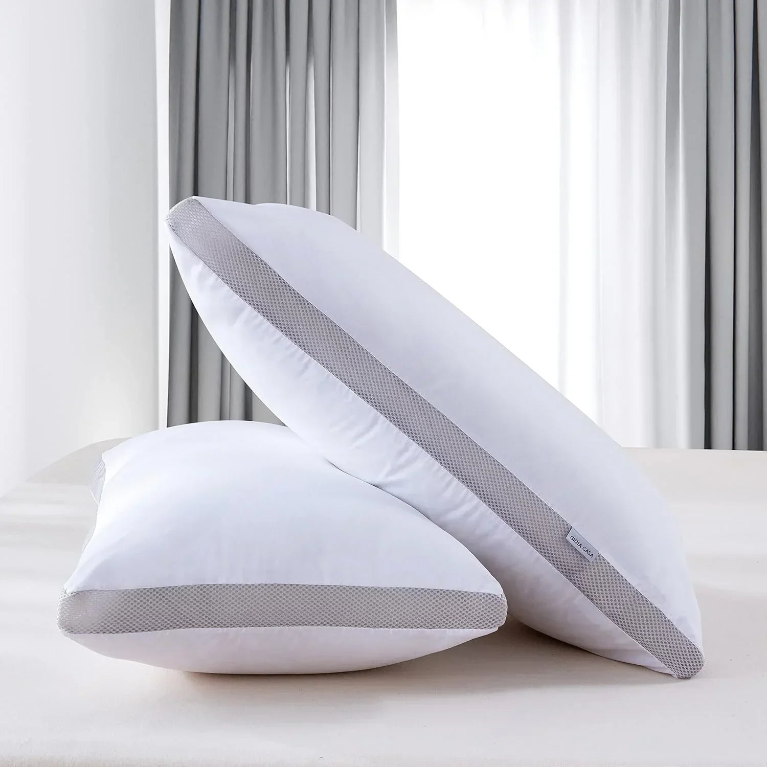 Two white pillows with gray mesh trim on a bed in a bright bedroom with gray curtains