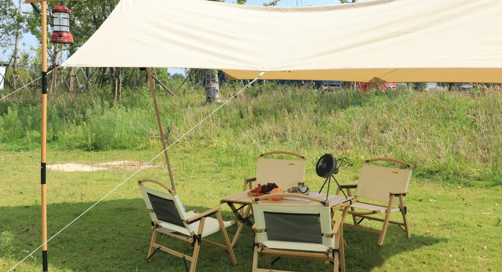 Outdoor camping setup with beige canopy, folding chairs, wooden table, lantern, and portable fan