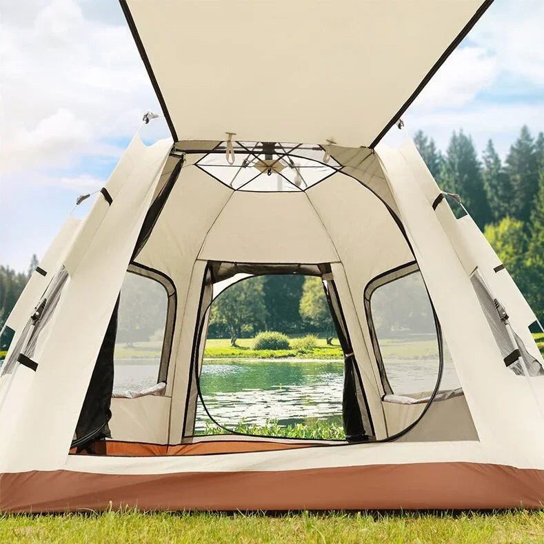 Open beige and brown camping tent set up on grassy lakeside with trees and water in background