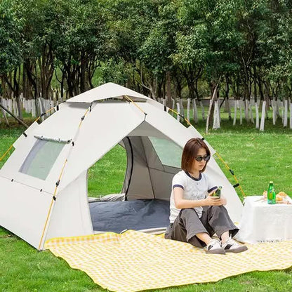Woman sitting on yellow checkered picnic blanket outside white camping tent in green park with trees